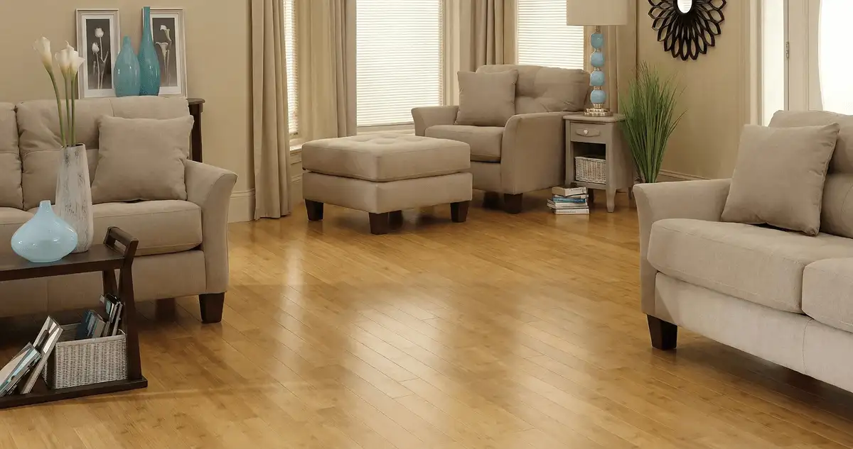 A living room with professionally installed bamboo flooring, beige sofas, a matching ottoman, a side table with a blue lamp, vases, framed pictures, and large windows with blinds and curtains in Sarasota, FL